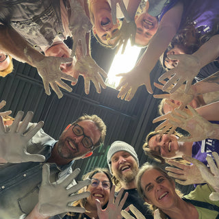 A circle of smiling potters seen from below, reaching pale clay-coated hands toward the camera at The Clay Hole in Draper, Utah
