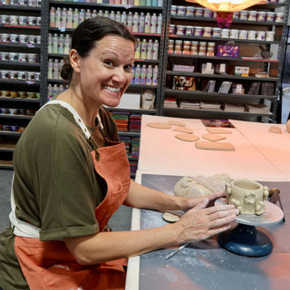 A smiling potter in a rust apron rests both hands on a clay cup with raised flowers on a turntable at The Clay Hole in Draper, Utah