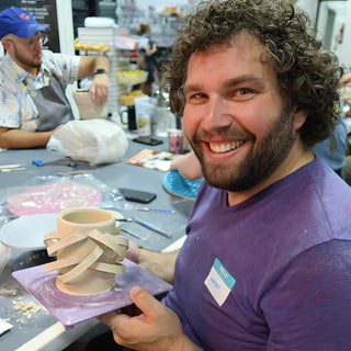 A grinning potter holding a small clay cup wrapped in crisscrossing flat bands of clay on a purple board at The Clay Hole in Draper, Utah
