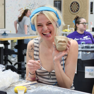 A potter in blue headphones sticking out their tongue while holding a small clay cup with sculpted lips at The Clay Hole in Draper, Utah