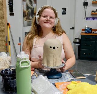 A potter wearing white headphones smiling behind a pale clay ghost with cut-out eyes and a triangle nose at The Clay Hole in Draper, Utah