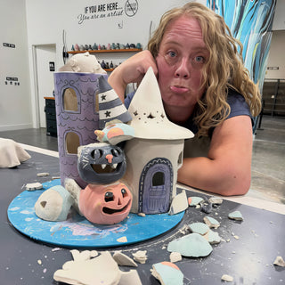 A potter with a pout leaning beside clay Halloween houses, a black cat and a pumpkin at The Clay Hole in Draper, Utah