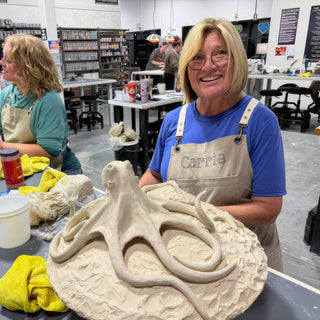 A smiling potter stands behind a large pale clay octopus with long curling arms on a textured round base at The Clay Hole in Draper, Utah