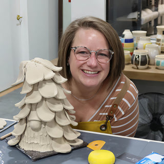 A smiling potter leaning in beside a tall clay sculpture covered in overlapping petal-shaped layers on a dark board at The Clay Hole in Draper, Utah