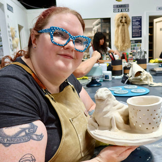 A potter holds up a round plate carrying an unglazed clay seal sculpture beside a cup with small round holes at The Clay Hole in Draper, Utah