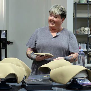 A smiling potter holding a flat tan object behind two large clay slabs draped over forms on turntables at The Clay Hole in Draper, Utah