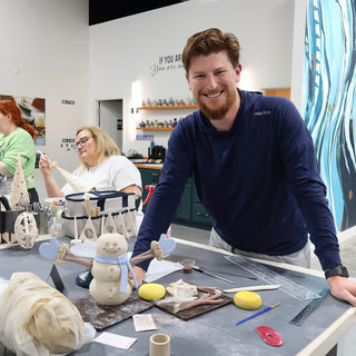 A smiling potter leans on a worktable beside a small clay snowman with a blue scarf and stick arms at The Clay Hole in Draper, Utah