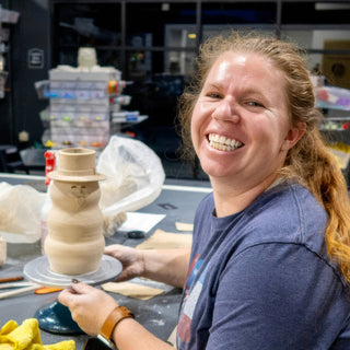 A smiling potter holds a turntable with a tan clay snowman wearing a top hat and a carved face at The Clay Hole in Draper, Utah