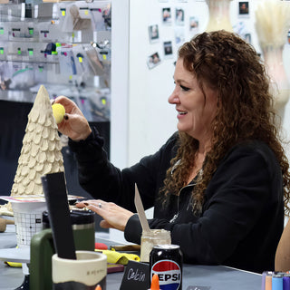 A smiling potter holding a yellow sponge beside a tall cone-shaped clay piece covered in overlapping scalloped layers at The Clay Hole in Draper, Utah