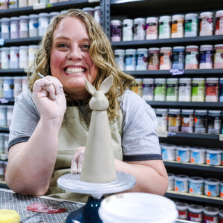 A grinning potter holding a small tool beside a tall, cone-shaped clay figure with bunny ears on a turntable at The Clay Hole in Draper, Utah