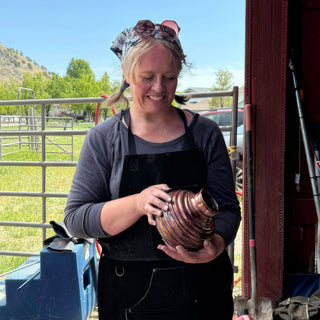 A potter in a patterned bandana smiles down at a small vase with a shiny copper finish and ribbed bands at The Clay Hole in Draper, Utah