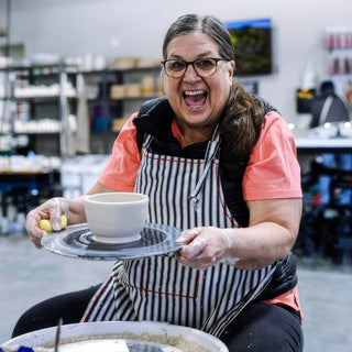 A laughing potter in a black and white striped apron holding up a cream-colored bowl sitting on a round board at The Clay Hole in Draper, Utah