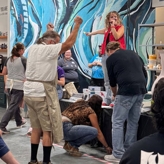 A person in a red vest holds a microphone on a table while a crowd cheers before a blue mural at The Clay Hole in Draper, Utah