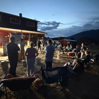 A crowd of people stands and sits in camp chairs outdoors at dusk facing a red building with mountains behind, with The Clay Hole, a pottery studio in Draper, Utah