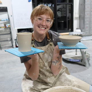 A smiling potter holding up a tall straight-sided cup and a small wide bowl, each on a blue board at The Clay Hole in Draper, Utah