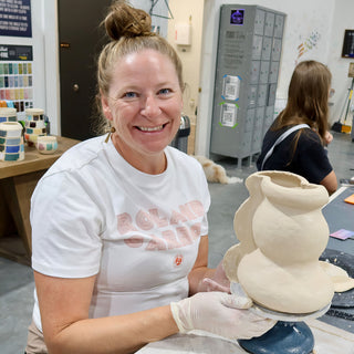A smiling potter in a clear glove supports a curvy cream clay vessel with two bulging sections on a turntable at The Clay Hole in Draper, Utah