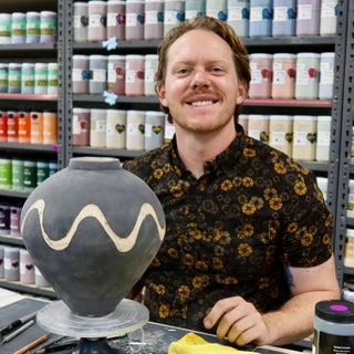 A smiling potter sits beside a rounded dark gray vase with a wavy cream stripe, shelves of glaze jars behind at The Clay Hole in Draper, Utah