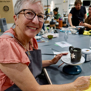A smiling potter sits at a table beside a dark gray mug with a white flower on a turntable at The Clay Hole in Draper, Utah