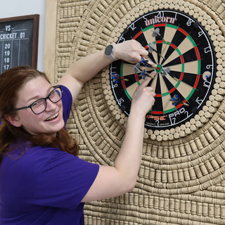 A smiling person in a purple shirt reaches for darts stuck in a dartboard ringed by wine corks at The Clay Hole in Draper, Utah