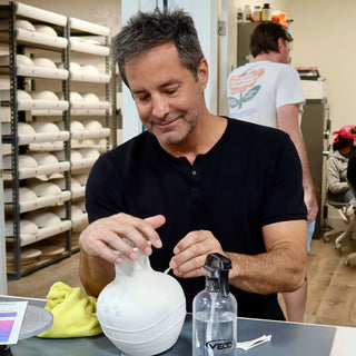 A potter smiles down at a white round vase with small blue dots, holding a fine tool against it at The Clay Hole in Draper, Utah