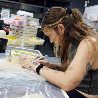 A potter with tattooed forearms bending over a table to work on a small clay cup with a tool at The Clay Hole in Draper, Utah