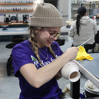 A potter in a knit beanie and purple t-shirt smiling while working a thin tool on a small white vase at The Clay Hole in Draper, Utah
