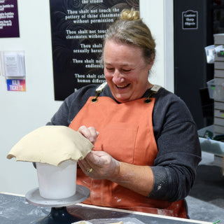 A smiling potter in an orange apron smoothing a wavy-edged clay slab draped over a white cylinder on a turntable at The Clay Hole in Draper, Utah