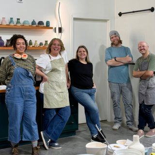 Five potters posing casually in aprons and overalls in front of a wall shelf of small colorful vases at The Clay Hole in Draper, Utah