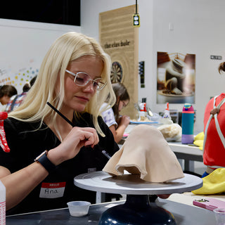 Wearing glasses, a potter works a thin tool along the folds of a tan clay form on a turntable at The Clay Hole in Draper, Utah