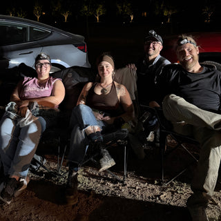 Four friends in headlamps sit close together in camp chairs and smile beside parked cars at The Clay Hole in Draper, Utah