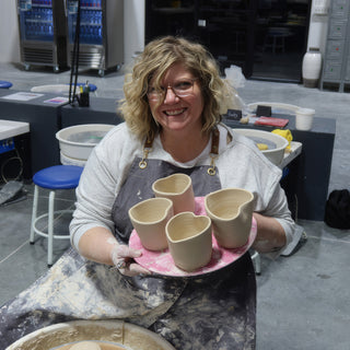 A smiling potter in a dusty gray apron holding a pink board with four unglazed cups pinched into heart shapes at The Clay Hole in Draper, Utah