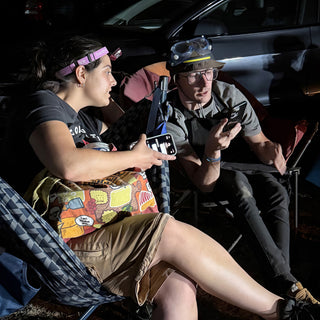 Two potters in headlamps and goggles sit in camp chairs and talk, one holding a phone at The Clay Hole in Draper, Utah