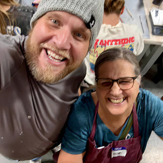 Two friends grinning in a selfie taken from above, one in a gray beanie and one in a maroon apron at The Clay Hole in Draper, Utah