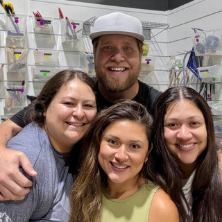 Four friends squeezed together in a smiling group selfie in front of a wall of hanging pottery tools at The Clay Hole in Draper, Utah