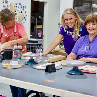 Three smiling people gather at a long gray table with a rolling pin, rolled clay and turntables at The Clay Hole in Draper, Utah