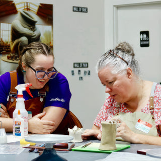 Two friends laughing together at a worktable, one working on a small textured clay mug with a looped handle at The Clay Hole in Draper, Utah