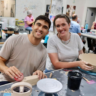 Two smiling people work at a table, one holding a bowl with a wavy rim, the other sponging clay at The Clay Hole in Draper, Utah