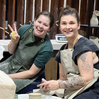 Two friends in aprons smiling at the camera, one holding up a peace sign with a yellow sponge in hand at The Clay Hole in Draper, Utah