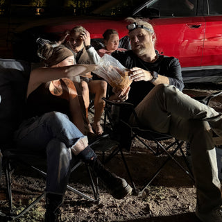 A group of friends relax in camp chairs in front of a red car, one reaching for a snack bag at The Clay Hole in Draper, Utah
