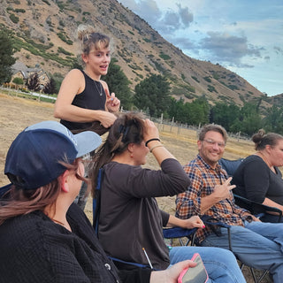 Friends sit in camp chairs on dry grass below a mountain hillside while a standing person talks at The Clay Hole in Draper, Utah