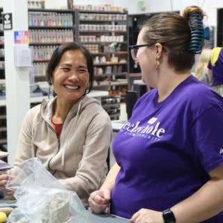Two friends laughing together at a work table with plastic-wrapped clay in front of them and shelves of glaze behind at The Clay Hole in Draper, Utah