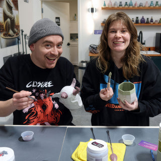 Two friends smiling at a table, one brushing a white sculpted piece, the other holding a pale green faceted cup at The Clay Hole in Draper, Utah