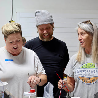 Three friends laughing over a worktable while two of them hold thin paintbrushes above small containers at The Clay Hole in Draper, Utah