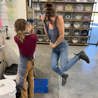 Two friends pulling on a long blue handle of a large machine while one kicks a boot up behind at The Clay Hole in Draper, Utah