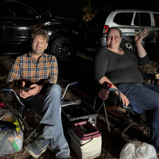 Two friends sit in camp chairs near parked cars, one waving at the camera, with a cooler beside them at The Clay Hole in Draper, Utah