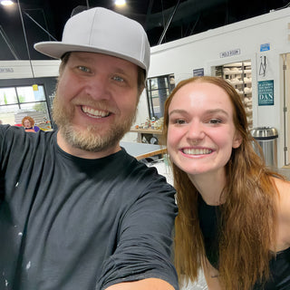 Two friends smiling in a close selfie inside the studio, one wearing a gray flat-brim cap and a dark t-shirt at The Clay Hole in Draper, Utah