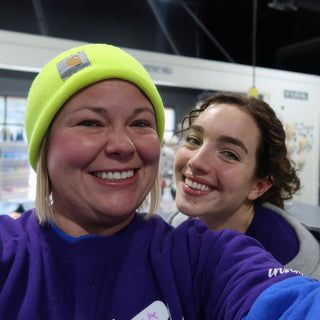 Two friends squeezed into a selfie, one in a bright yellow-green beanie, both in purple shirts and smiling at The Clay Hole in Draper, Utah