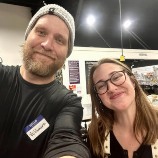 Two friends smiling in a selfie, one in a gray beanie with a name tag and one wearing glasses at The Clay Hole in Draper, Utah