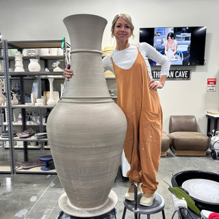 A potter in orange overalls stands on a stool beside a very tall unglazed gray vase with a flared neck at The Clay Hole in Draper, Utah