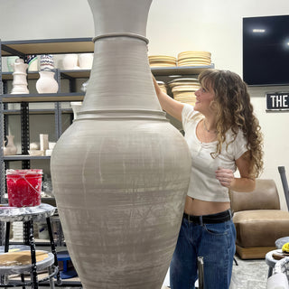 A smiling potter resting a hand on a giant two-part unglazed vase with a narrow neck that towers over them at The Clay Hole in Draper, Utah